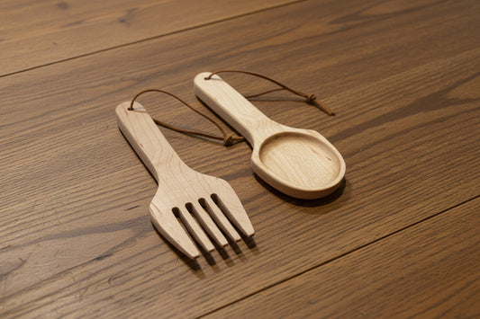 Two small wooden utensils with brown hanging cords, placed on top of printed white sheets. One utensil has a round bowl-like head, and the other has a fork-like head with several tines.